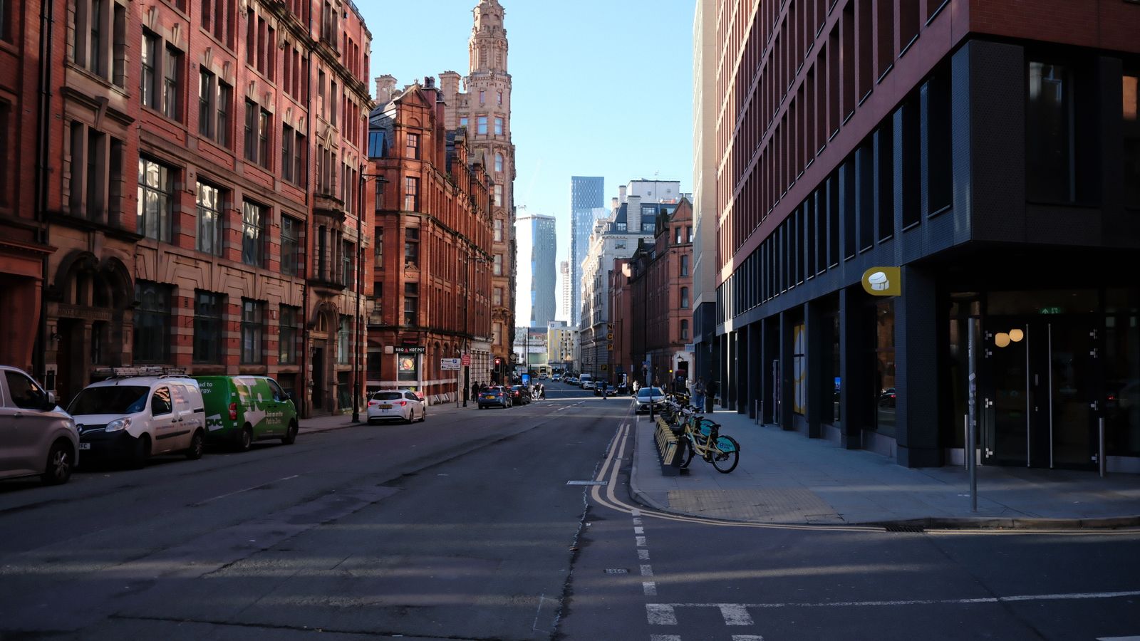 A Manchester street lined with red-brick Victorian buildings, parked cars, and hire bikes, with modern glass towers visible in the distance.