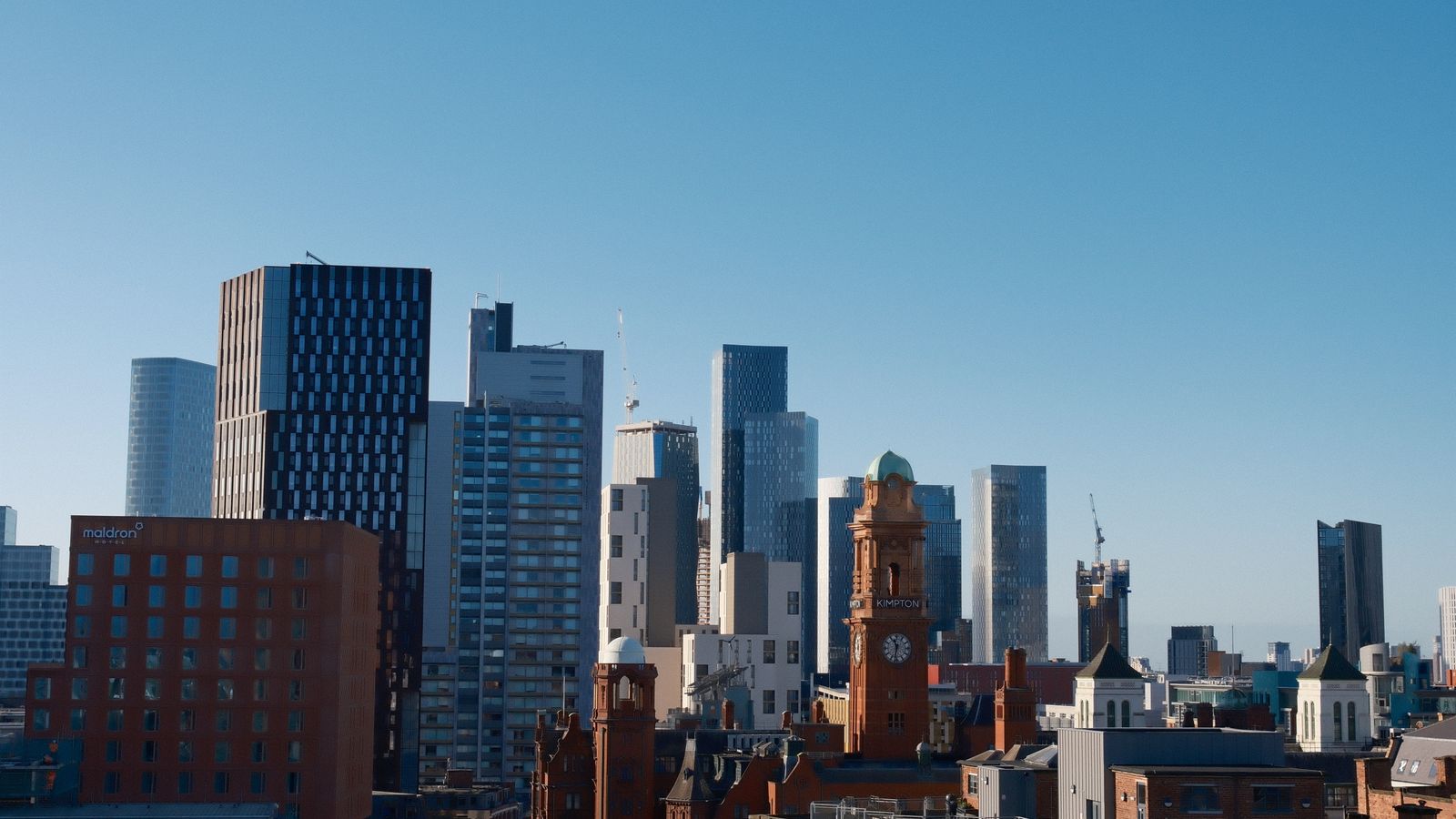 Manchester city skyline on a clear day, showing modern glass towers, the red-brick Maldron Hotel, and a Victorian clock tower among high-rise buildings.