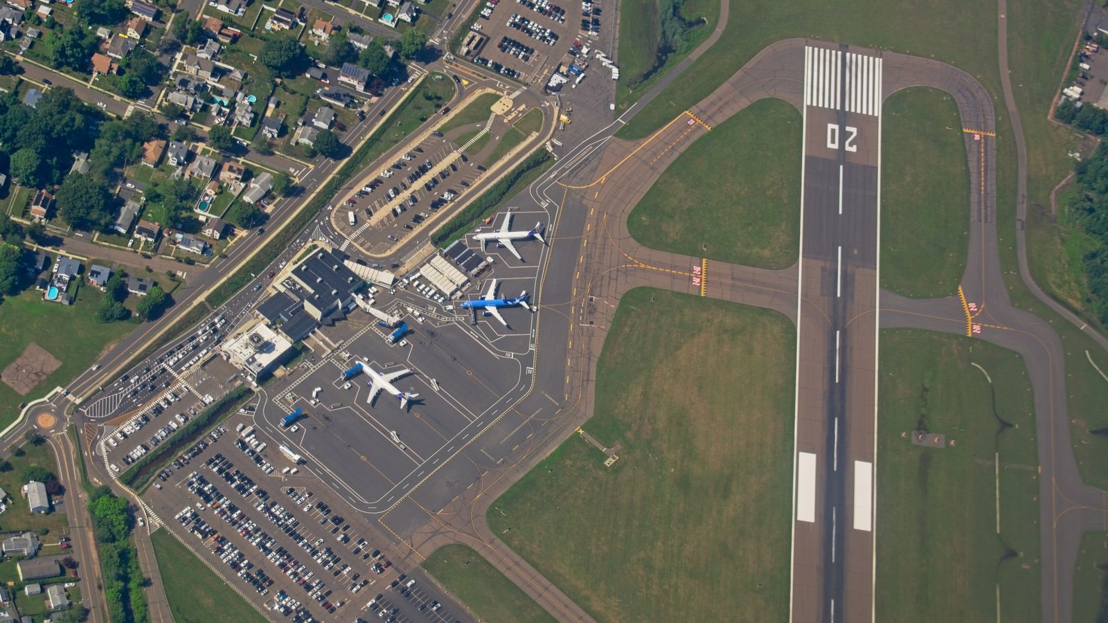 Aerial view of an airport runway and terminal, showing parked aircraft, taxiways, and surrounding car parks and streets.